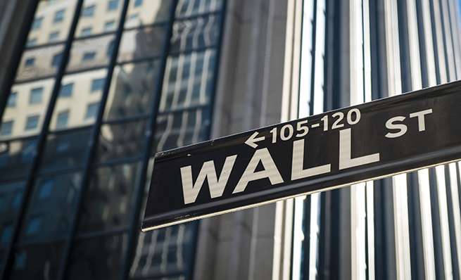 A close-up of a black street sign that reads "WALL ST" with an arrow, set against the backdrop of reflective skyscraper windows and a fluted column.