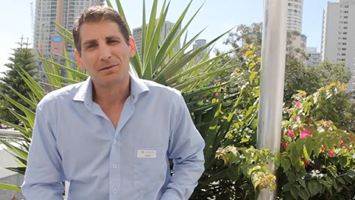 Man in blue shirt outdoors with tropical plants and city skyscrapers behind him.v