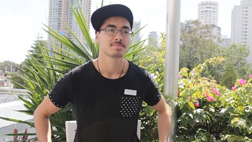 Young man with glasses and backward cap in black polka-dot shirt outdoors by city buildings.