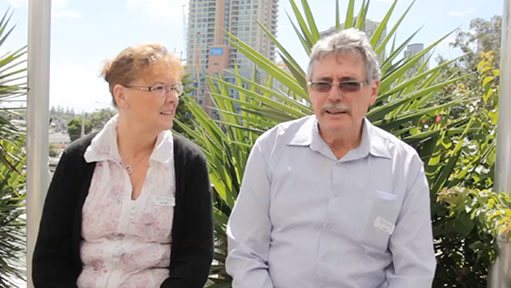 Older couple in glasses seated outdoors with tropical plants and city high-rises behind them.