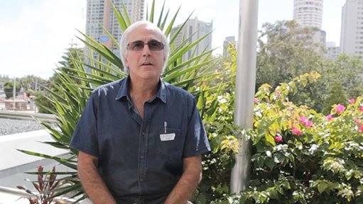 Older man in sunglasses and dark blue shirt standing outdoors by tropical plants and city buildings.