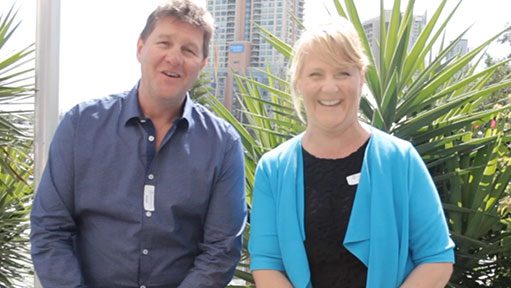 Man in blue shirt and smiling woman in blue cardigan outdoors by palm plants and city buildings.