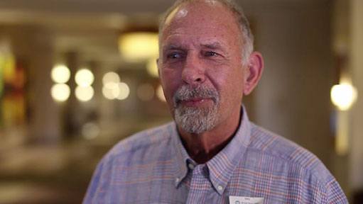 Older man with goatee and plaid shirt looking thoughtful in a warm indoor setting.