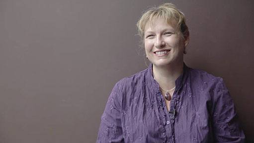 Blonde woman named Keetha in a purple wrinkled shirt smiling warmly against a dark background.