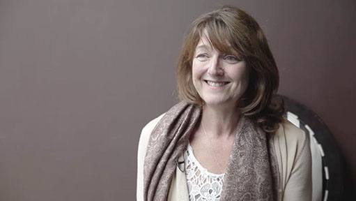 Smiling woman with bangs and a brown scarf against a plain reddish-brown background.