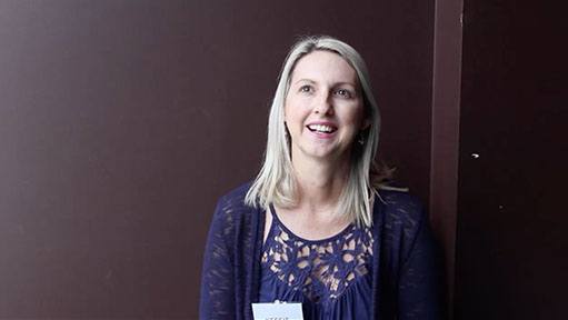 Smiling blonde woman in a dark blue top and cardigan against a brown wall.
