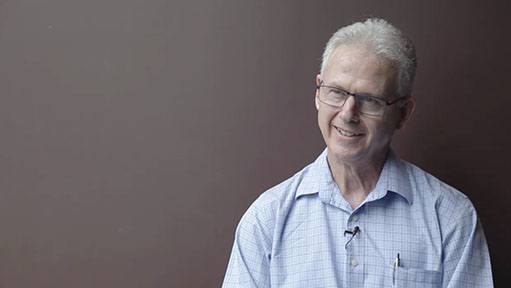 Smiling man with white hair, glasses, and a blue checkered shirt against a brown background.