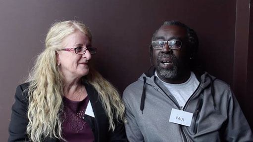 Woman and man in glasses seated together against a brown background, man wearing "PAUL" tag.