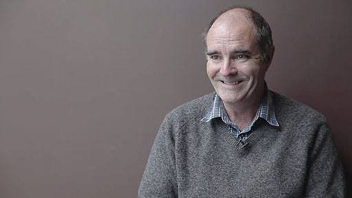 Older man with grey sweater and collared shirt smiling against a plain brown background.