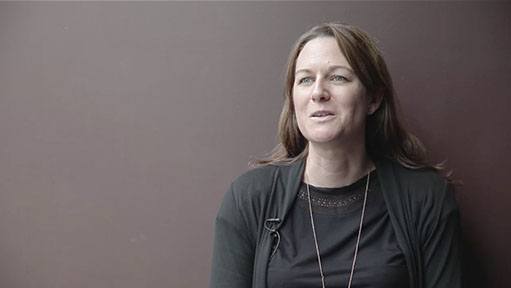 Woman in black top and cardigan against a brown background, looking thoughtful.