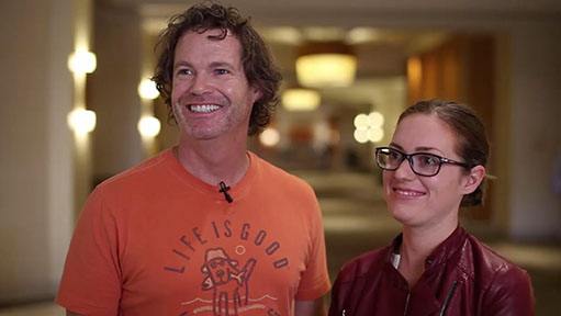 Man in "LIFE IS GOOD" shirt and woman in red jacket and glasses smiling indoors.