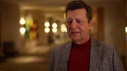 Man in grey blazer and red turtleneck standing indoors with a warm, blurred background.
