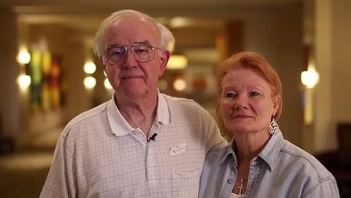 Older man in glasses and a red-haired woman with large earrings standing together indoors.
