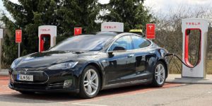 A black Tesla electric vehicle parked at a Supercharger station with charging stalls visible.