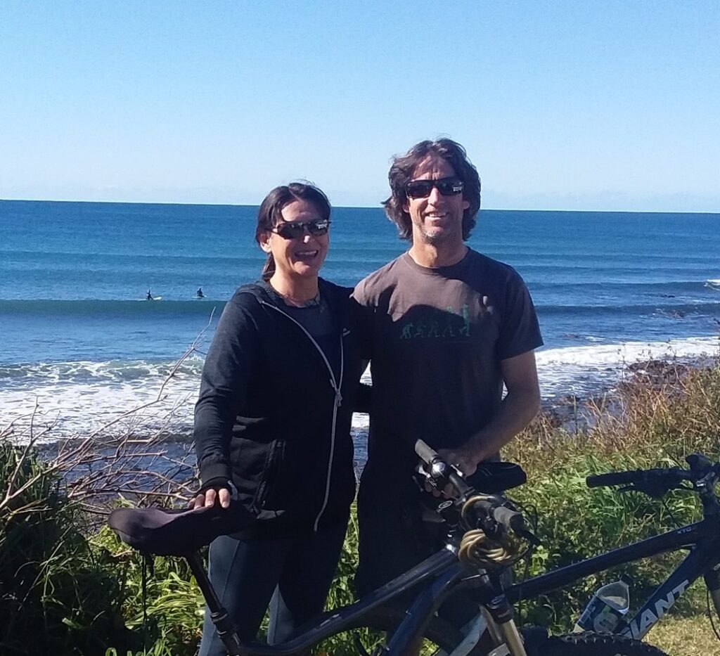 A smiling man and woman wearing sunglasses and casual clothing stand with their mountain bikes near a grassy cliff overlooking a sunny blue ocean with surfers.