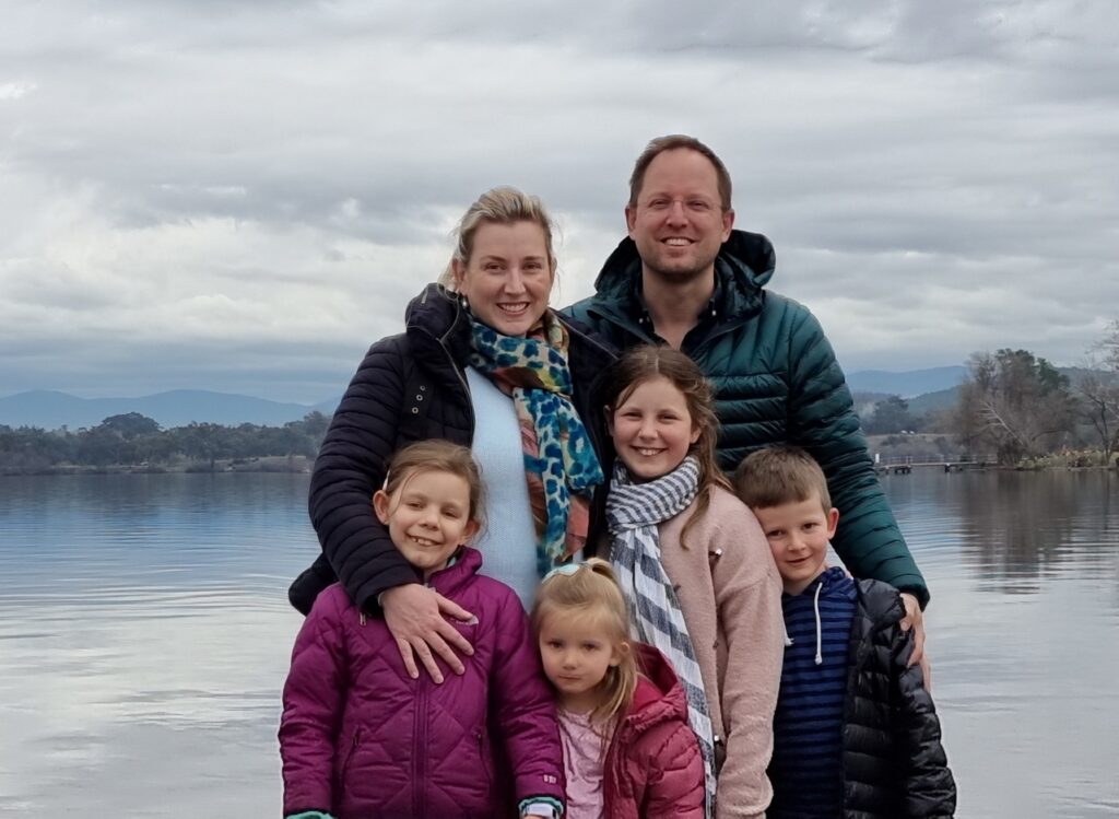 A smiling family of six, two adults and four children, stand bundled up in jackets and scarves by a calm body of water with mountains in the distance under a cloudy sky.