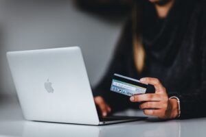 A close-up, low-light photo of a person's hands holding a debit or credit card near the keyboard of a silver Apple laptop, signifying an online transaction or e-commerce.