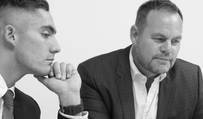 A black and white close-up of two men in suits, one younger and one older, both with serious expressions, looking down and concentrating on something out of frame.