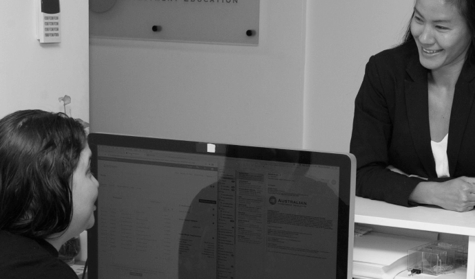 A black and white photograph of two women smiling while one sits at a computer reviewing information and the other stands at a counter, possibly discussing a screen on the monitor.
