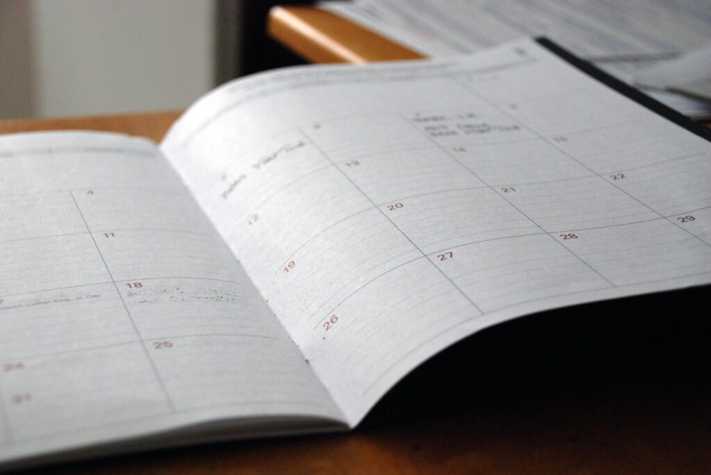 A close-up shot of an open paper planner or calendar on a wooden desk, showing blank monthly grids with faint handwritten notes.