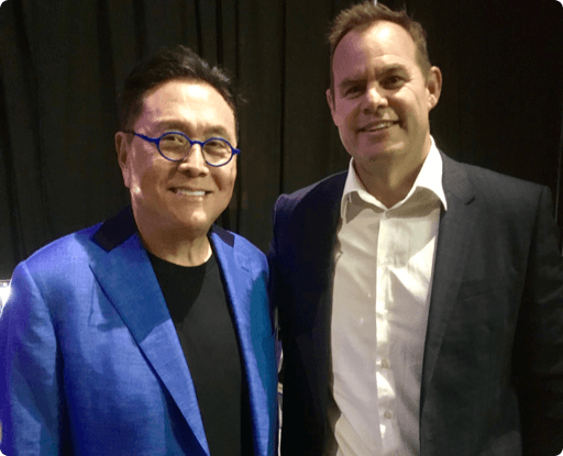 A photo of two men, Robert Kiyosaki in a bright blue jacket and glasses, smiling next to a Caucasian man in a dark suit jacket and white shirt.