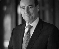 A black and white portrait of a middle-aged man in a dark, patterned suit and patterned necktie, looking directly at the camera.