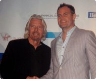 A photo of two men, one resembling Richard Branson with long white hair in a black shirt, posing next to a younger man in a light-colored striped suit jacket, at a financial event.