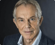 A close-up portrait of former UK Prime Minister Tony Blair, with grey hair, wearing a dark jacket and a light blue shirt, against a muted background.
