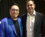 A photo of two men: Robert Kiyosaki, wearing a bright blue jacket and blue-framed glasses, smiling next to a Caucasian man in a dark suit jacket and white shirt.