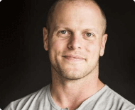 A close-up portrait of a smiling bald Caucasian man with a slight beard, wearing a light grey t-shirt against a dark background.